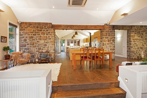 A warm open-plan interior combining a dining area, kitchen, and living space. Exposed rough stone walls, a substantial timber beam, polished dark timber flooring, and white plaster ceilings create a rustic yet refined character. The dining table sits centrally beneath pendant lights, with a kitchen beyond and a small sitting area to the left. Steps and low white built-in cabinetry frame the foreground.
