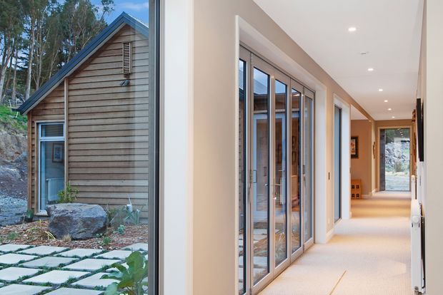 Split-view image showing a compact timber-clad gabled cabin exterior on the left, set among rocky natural landscaping, and a bright contemporary interior hallway on the right. The hallway features floor-to-ceiling glazed doors or windows, white walls, light carpet, recessed downlights, and warm timber accents, with views toward the surrounding landscape.