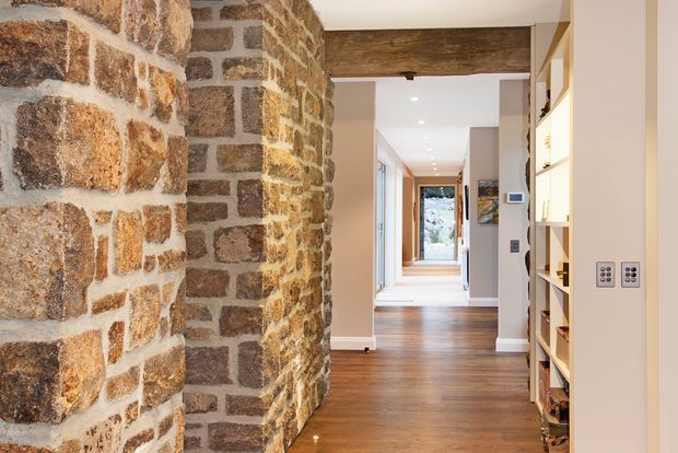 Interior view down a long hallway with a prominent exposed rough-stone masonry wall and a dark timber beam overhead. The corridor has warm timber flooring, light painted walls, recessed ceiling lights, built-in open shelving on the right, and framed artwork. A glazed opening at the far end brings in daylight and views outside.