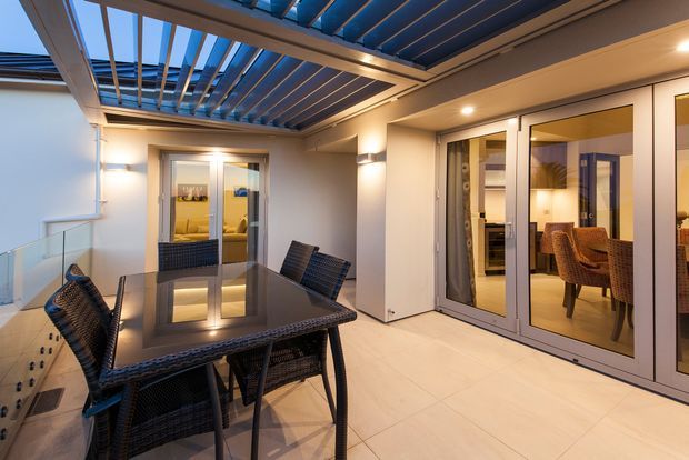 A contemporary covered outdoor dining terrace with a glass-topped dark woven dining table and chairs. The terrace has pale tiled flooring, glazed sliding doors opening into a warmly lit interior dining area, and a striking overhead louvred pergola with blue-toned slats and integrated lighting. Glass balustrades and clean white walls create an airy, refined setting.