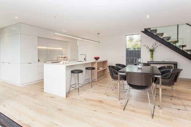 Bright open-plan contemporary kitchen and dining area with white handleless cabinetry, a central island with bar stools, pale timber flooring, and a dark dining table with black chairs. A glazed stair balustrade and staircase are visible at the right, along with integrated shelving and large windows.