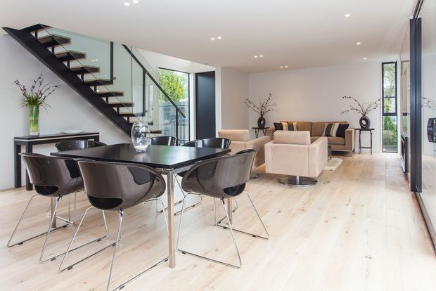 Bright open-plan contemporary interior combining a dining area and living room. The space features pale timber flooring, white walls, extensive glazing, a sculptural black open-riser staircase with glass balustrade, a dark rectangular dining table with modern black chairs, and a neutral-toned lounge area.