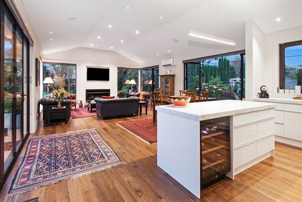Open-plan contemporary kitchen and living area with warm timber flooring, a large white island with integrated wine storage, extensive black-framed glazing, and a furnished lounge with fireplace and television. The room is bright and connected to a landscaped garden through the windows.