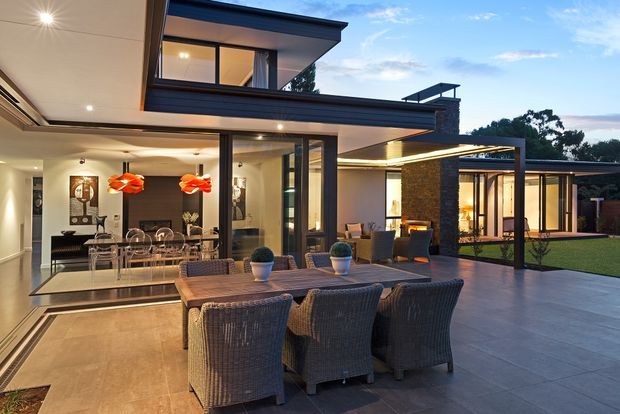 Twilight view of a sophisticated contemporary home with extensive floor-to-ceiling glazing, flat rooflines, dark metal framing, and a large tiled outdoor entertaining terrace. A dining table with woven chairs occupies the foreground, while the open-plan interior, illuminated kitchen and dining area, covered outdoor lounge, landscaped lawn, and stone-clad feature wall are visible beyond.