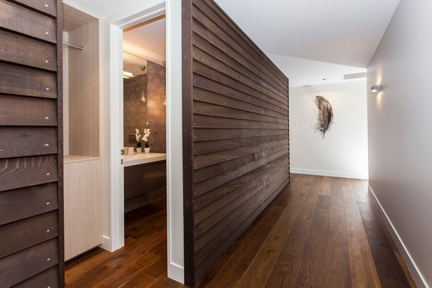 A refined residential hallway with dark timber flooring and a striking full-height horizontal timber feature wall enclosing a bathroom doorway. White walls and ceiling create contrast, while pale timber built-in cabinetry frames the open bathroom entrance. The bathroom is visible through the doorway with a stone-look wall finish, vanity, and decorative styling. A sculptural wall feature and discreet wall lighting complete the corridor.