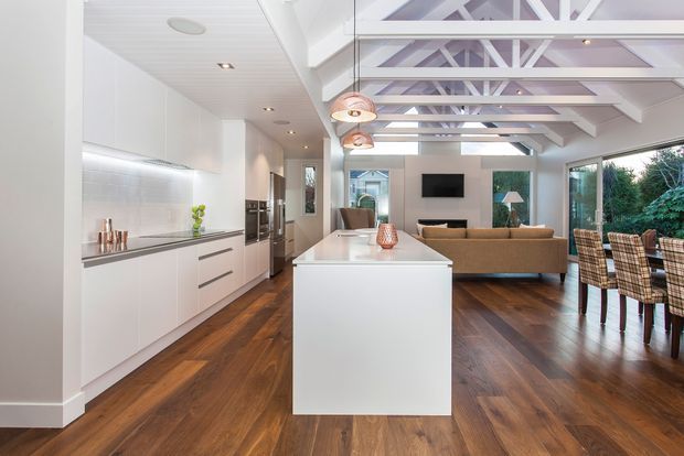 Bright open-plan kitchen and living area with white handleless cabinetry, a large central island, warm timber flooring, exposed white roof trusses, pendant lights, and extensive glazing opening toward a leafy outdoor setting. A dining area and living room are visible beyond the kitchen.
