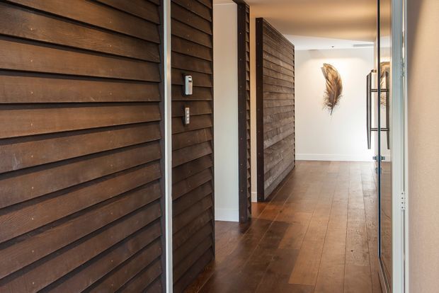 A warm, modern entry hallway viewed from a glazed doorway. The space features dark horizontal timber wall cladding, rich timber flooring, white plaster walls, dark door frames, and a minimalist wall-mounted feather-like artwork. A narrow black metal shelving unit is visible at the far end.