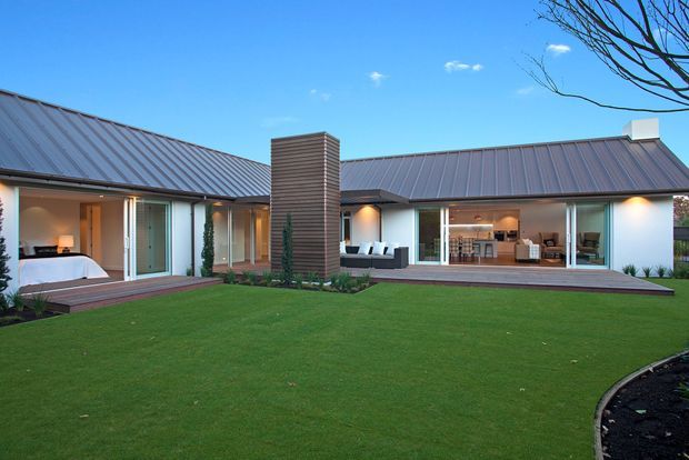 Single-storey contemporary house viewed from a landscaped courtyard, with dark standing-seam metal gabled roofs, extensive sliding glass doors, timber decking, and a prominent vertically clad central chimney or feature wall. Bedroom and living/dining interiors are visible through the glazing.