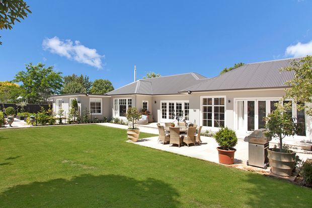 A single-storey residential home viewed from a landscaped lawn and courtyard-like outdoor area. The house has pale rendered walls, a large grey hipped metal roof, extensive white-framed French doors and windows, and a paved patio furnished with an outdoor dining setting and potted plants. The scene is bright and sunny with a calm, formal garden setting.