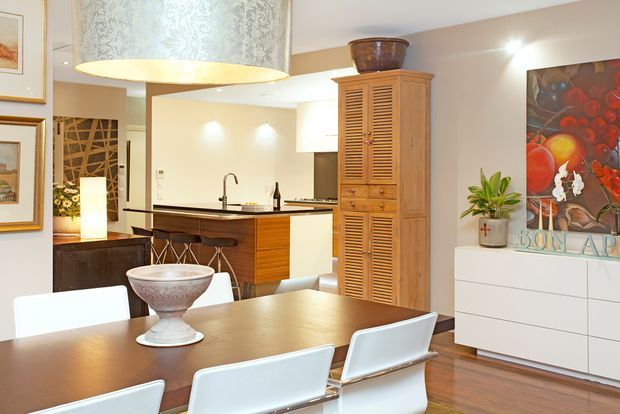 A warm, polished open-plan dining and kitchen interior. A large wooden dining table with white chairs occupies the foreground, while the background features a compact kitchen with dark benchtops, timber cabinetry, a sink and island stools. A tall louvred timber storage cabinet, white sideboard, artwork, indoor plants and a large decorative pendant light complete the space.