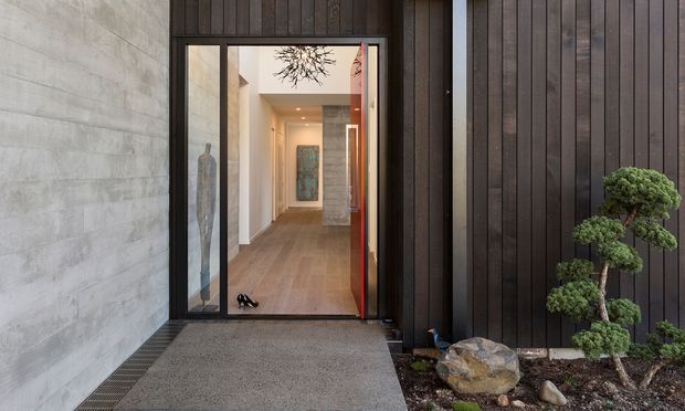 Contemporary residential entrance with a dark vertical timber-clad facade, exposed concrete wall, black-framed glazed pivot door, and a sheltered concrete entry path. Through the doorway, a warm timber-floored hallway with a sculptural pendant light and artwork is visible. A carefully pruned ornamental tree and stone landscaping sit beside the entrance.
