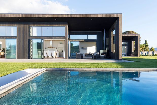 A contemporary single-storey home with strong horizontal and rectilinear forms, dark timber-toned cladding, expansive floor-to-ceiling glazing, and a deep covered outdoor living area. The house overlooks a turquoise swimming pool set within a manicured lawn, with a visible dining area and lounge furnishings inside and beneath the covered terrace.