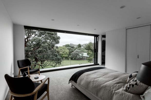 A contemporary bedroom with a low-profile upholstered bed, dark bedding, black lounge chairs with timber frames, and a small round side table. Full-height glazing provides expansive views of mature trees and a green suburban landscape. The room has a restrained monochrome palette, recessed ceiling lights, built-in white wardrobe cabinetry, and a soft carpeted floor.