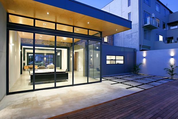 Contemporary urban residential courtyard viewed from outside, featuring a dark blue rendered facade, expansive black-framed floor-to-ceiling glazing, and a projecting timber-lined canopy with recessed downlights. The glazed interior reveals a minimalist living area with a dark island or built-in bench, while the courtyard combines a concrete terrace, timber decking, large square pavers, and integrated wall lighting.