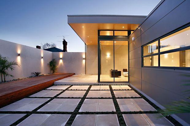 Contemporary residential courtyard viewed from outdoors at dusk, featuring a dramatic flat-roofed entry volume with warm recessed lighting, full-height glazing, dark metal framing, and a horizontal grey-clad facade. Large pale square pavers set in a regular grid with grass joints lead toward the glazed entrance, while a raised timber deck and minimalist planted courtyard border the space.
