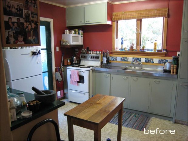 A compact older kitchen shown in a “before” renovation state, with red painted walls, pale green-grey cabinetry, a freestanding white stove, stainless-steel sink and benchtop, patterned tile splashback, refrigerator, microwave, and a central timber work table. The room has warm natural light, a traditional utilitarian layout, and visible family photo displays.