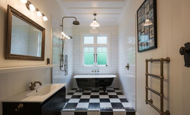 A bright, narrow traditional bathroom with white subway-tiled walls, a black-and-white checkerboard tiled floor, a freestanding clawfoot bathtub beneath a sash window, a wall-mounted shower, a vanity with basin, an ornate framed mirror, and a brass heated towel rail. The room features decorative ceiling panel moulding and classic brass fixtures.
