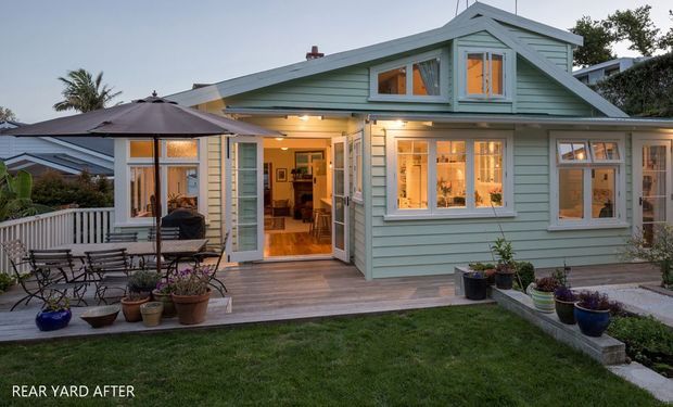 Rear-yard view of a compact, light-green weatherboard cottage with a pitched gable roof, white-trimmed windows, and warm interior lighting visible through the glazing and open rear door. A timber deck, outdoor dining furniture, large umbrella, potted plants, lawn, and low garden edging form the outdoor setting. The image is captioned “REAR YARD AFTER.”