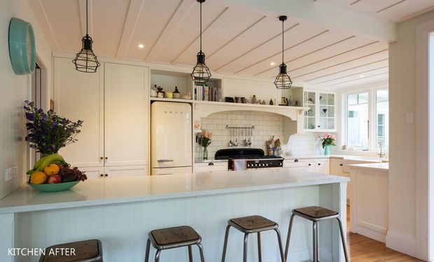Bright, spacious traditional kitchen with a large pale island, integrated cabinetry, white brick-tile splashback, open shelving, and a range cooker. The room features a white beadboard-style ceiling with exposed linear detailing and black cage pendant lights over the island. Timber flooring, pale cabinetry, and abundant daylight create a warm, inviting atmosphere.