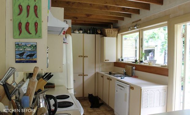 Compact, traditional kitchen with cream built-in cabinetry, a white freestanding stove and dishwasher, timber benchtops, tiled flooring, exposed timber ceiling beams, and broad windows providing abundant natural light. Cooking utensils and vintage-style decorative details contribute to a lived-in country character.