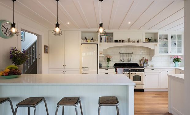 A bright, welcoming traditional-style kitchen viewed from the island. The room features white shaker-style cabinetry, a large pale island with three stools, a freestanding retro-style refrigerator, a range cooker beneath a white subway-tile backsplash, glass-front upper cabinets, timber flooring, and a white tongue-and-groove panelled ceiling. Black cage pendant lights provide contrast, while a stairway and entry area are visible at the left.