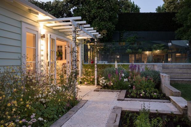 A landscaped courtyard and garden walkway beside a light-coloured weatherboard building, viewed at dusk. The path passes through abundant planted garden beds and beneath a white timber pergola, with warm lighting illuminating glazed doors and windows. Raised concrete or rendered garden edges and steps lead toward a darker adjoining building in the background.