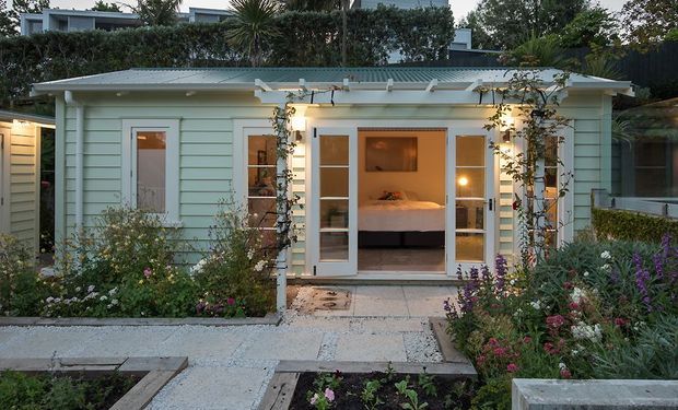 A compact single-storey garden dwelling with pale blue horizontal weatherboard cladding, a shallow corrugated metal roof and glazed French doors opening onto a paved path. Flowering garden beds and climbing plants frame the facade, while a bedroom with a bed is visible through the open doors. The setting has a calm, landscaped residential character.
