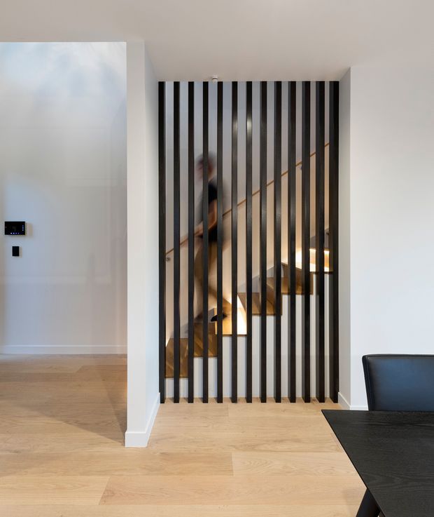 A bright, minimalist interior showing a black vertical slatted screen separating the entry area from a staircase. The staircase has timber treads and a dark handrail, with warm integrated lighting beneath or alongside the steps. Pale timber flooring, white walls, and a partial black dining table and chair create a clean contemporary scheme.
