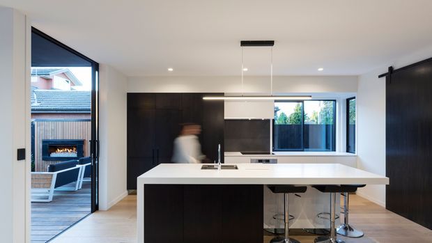 Contemporary open-plan kitchen with a large white island and integrated seating, dark timber cabinetry, light flooring, and a slim linear pendant. Full-height glazing and an open sliding door connect the kitchen to an outdoor deck with a built-in fireplace.