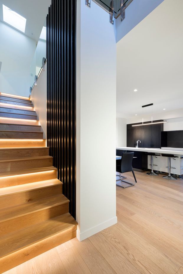 A contemporary open-plan interior featuring a timber staircase with warm LED strip lighting beneath each tread, a tall black vertical slat feature beside the stairs, and a modern kitchen visible beyond. The kitchen has dark cabinetry, a white island with bar seating, integrated pendant lighting, pale timber flooring, and crisp white walls. A partial upper-level void or landing is visible above.