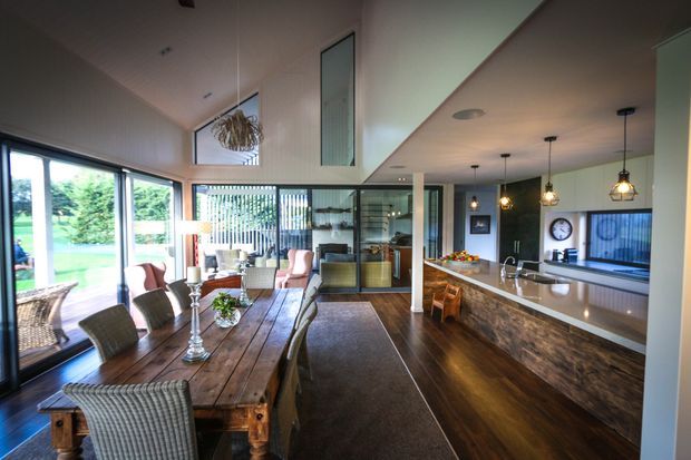 Open-plan interior combining a dining area, living space and kitchen. A large rustic timber dining table occupies the foreground, with woven upholstered chairs and a long runner. The kitchen features a substantial island with a dark stone-look benchtop, timber-faced cabinetry, pendant lights and integrated appliances. Floor-to-ceiling glazing opens toward a green outdoor area, while the room has dark timber flooring, pale walls, high ceilings and a contemporary open layout.