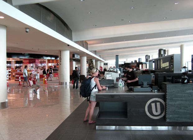 A bright, spacious commercial interior resembling an airport or transport terminal, with a dark stone service or café counter in the foreground, staff and customers, polished floors, tall white columns, layered ceiling planes, extensive glazing, and retail concessions in the background. The counter features a prominent circular logo, while the broad open circulation area and exposed structural ceiling create a modern public-space atmosphere.