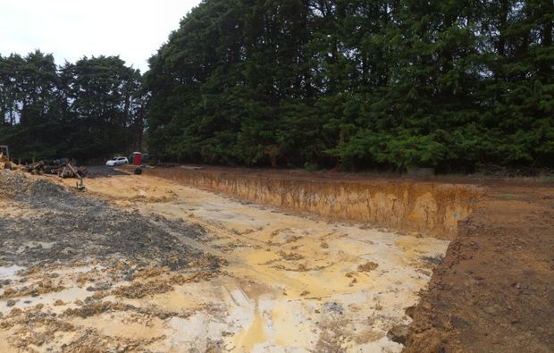 A cleared and excavated construction site with exposed sandy soil, muddy ground, a cut earth bank, and dense mature trees along the rear boundary. Construction equipment and vehicles are visible in the distance.