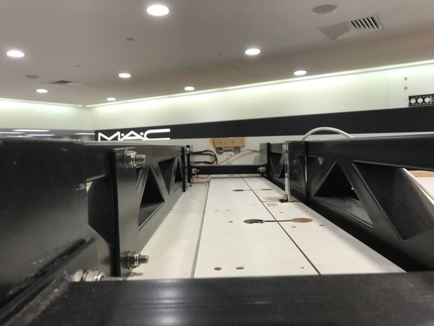 Close-up view of a large-format commercial interior installation, showing black structural or equipment housings flanking a central white panelled surface. The space has a low suspended ceiling with recessed lighting, visible ventilation grilles, exposed cabling and technical hardware, and a black MAC-branded wall element in the background.