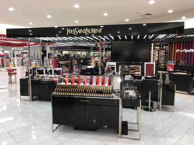 A high-end department-store cosmetics and beauty retail counter dominated by a black Yves Saint Laurent branded display. The open commercial space features glossy tiled flooring, mirrored and black display cabinetry, illuminated product shelving, makeup testers, counters, stools, and bright recessed ceiling lighting. Red and pink accents appear throughout the branded retail fit-out.