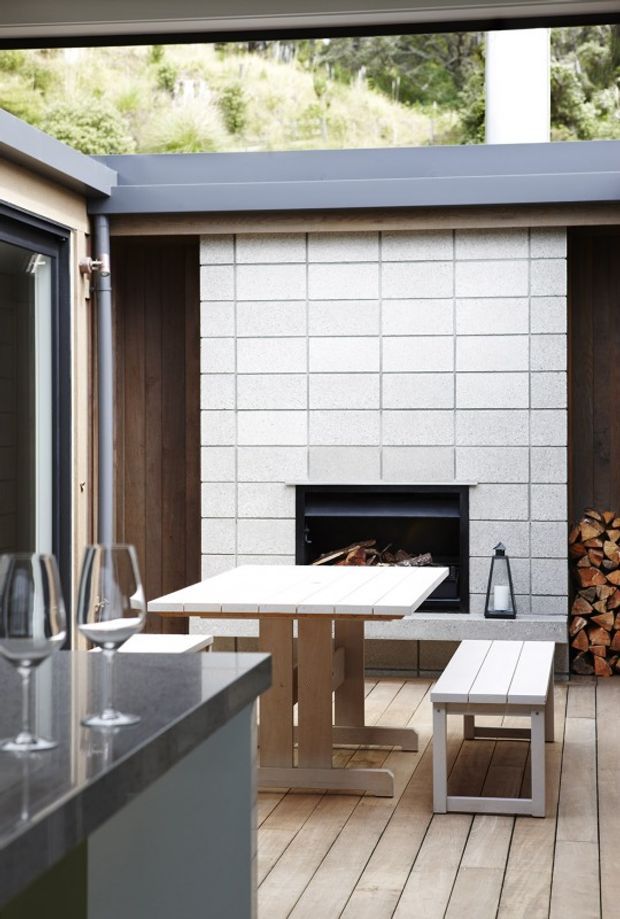 A contemporary covered outdoor entertaining area viewed from an adjoining kitchen. The space features a white rectangular tiled fireplace surround with a built-in firebox, dark timber wall lining, a grey metal roof edge, white outdoor dining table and bench seating, stacked firewood, and timber-look decking. A dark stone kitchen benchtop and wine glasses are visible in the foreground.