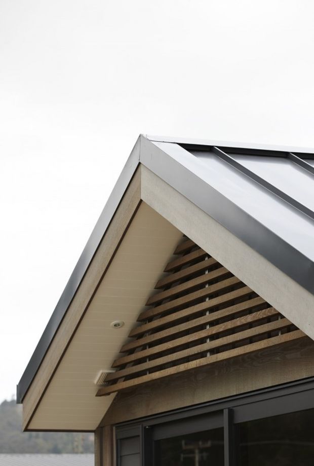 Close-up upward view of a contemporary gabled building facade, showing a sharply pitched roof with dark metal standing-seam roofing, light wood-toned soffits and cladding, exposed horizontal timber screening beneath the gable, and dark-framed glazing at the lower edge.