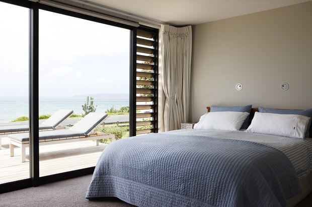 A bright coastal bedroom with a large black-framed glazed sliding door opening onto a timber deck and ocean view. The room contains a neatly made bed with blue-grey textured bedding, white pillows, neutral walls, and full-height curtains. Outdoor loungers are visible beyond the glazing.