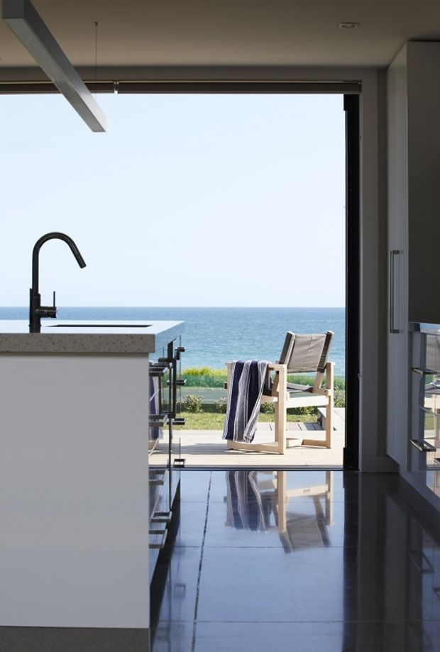 A sleek modern kitchen interior opens directly onto a sunlit deck overlooking the ocean. The space features a white island with a black gooseneck tap, dark glossy tiled flooring, minimalist cabinetry, broad glazed doors, and a simple outdoor chair with a towel on the deck. The strong indoor-outdoor connection and ocean horizon create a bright coastal setting.