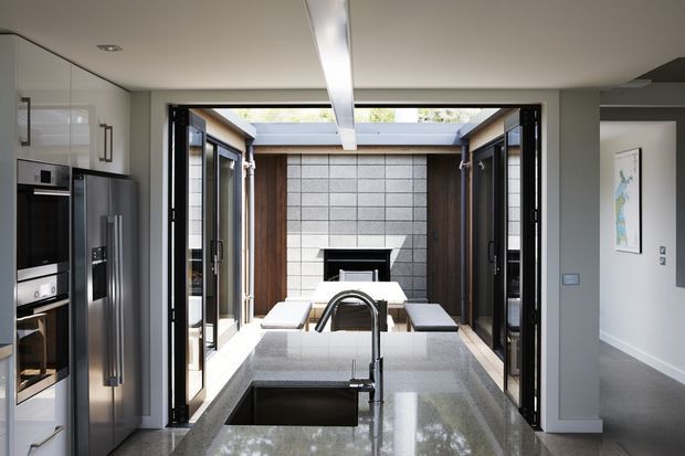 A contemporary kitchen viewed toward an open courtyard or outdoor entertaining area. The room features white full-height cabinetry with integrated stainless-steel appliances, a large polished grey stone island with undermount sink and curved black tap, and expansive black-framed folding or sliding doors opening to a sheltered outdoor space. The courtyard includes dark timber-clad walls, grey masonry feature blocks, a fireplace or barbecue-like opening, and built-in bench seating. The kitchen has a clean, bright, highly connected indoor-outdoor layout.