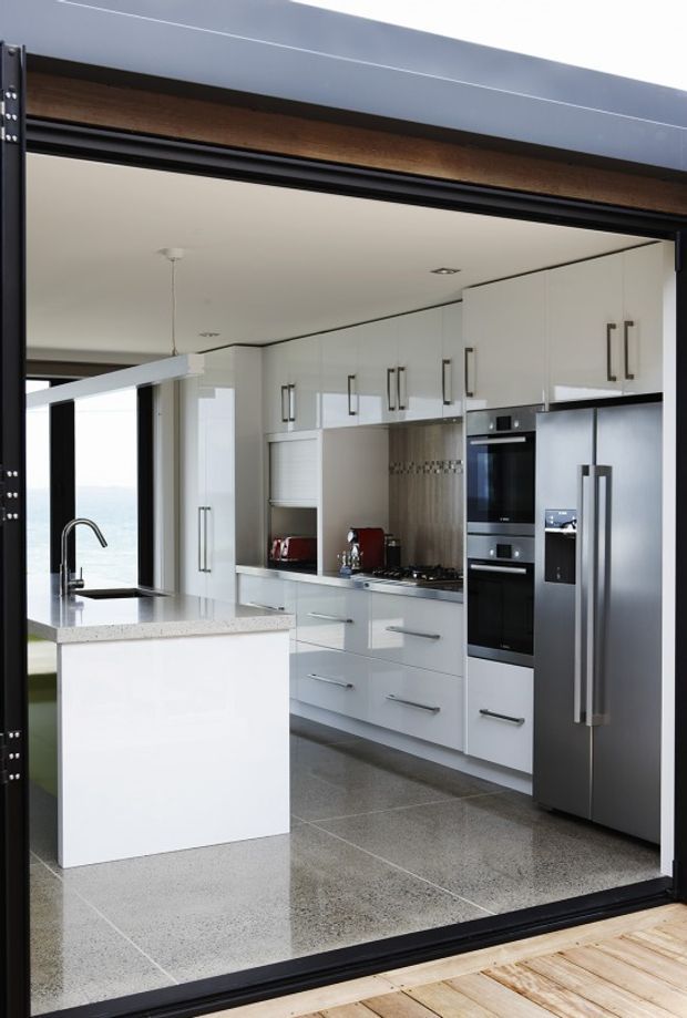 A bright contemporary kitchen viewed through a large black-framed opening. The room features glossy white full-height cabinetry with long dark handles, integrated double ovens, a stainless-steel refrigerator, a dark stone or composite benchtop, and a compact island with an undermount sink and tall curved tap. Large grey polished floor tiles and a textured neutral splashback complete the clean, modern fit-out. Timber decking is visible outside the opening, with a partial water view beyond.