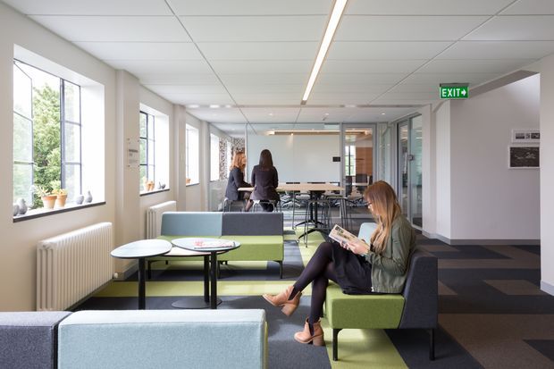 A bright contemporary commercial interior, likely a school, library, or office common area, with large windows, lounge seating, small round tables, a communal table with chairs, and a corridor leading toward an exit. The space has a suspended acoustic tile ceiling, linear recessed lighting, carpeted flooring, and a restrained palette of white, grey, black, and muted green.