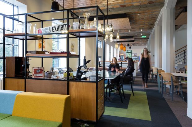 An open-plan commercial interior with exposed brick and timber ceilings, black-framed glazed and open shelving used as a room divider, contemporary office seating, workstations, and a casual dining or breakout area. The space combines industrial architectural features with colourful upholstered furniture, patterned carpet tiles, pendant lighting, and visible stair access.