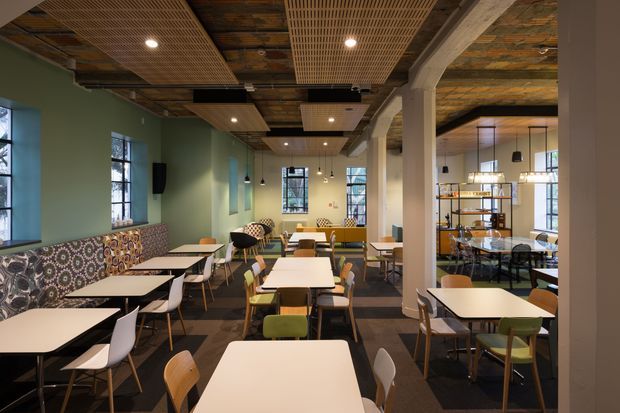 A contemporary commercial dining space or café with numerous light rectangular tables and mixed chairs, a long patterned banquette along a muted green wall, large black-framed windows, pendant lighting, and exposed timber ceiling elements. Structural columns divide the room, while a glazed shelving or service area is visible at the rear. The interior combines warm timber, dark flooring, patterned upholstery, and soft green walls.