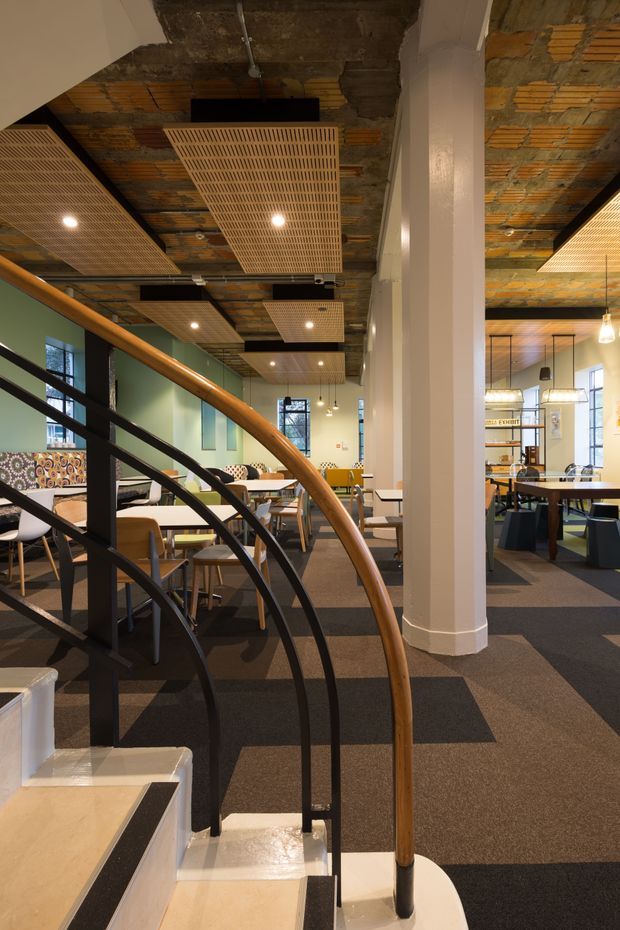 A contemporary commercial interior, likely a café, restaurant, or hospitality dining space, viewed from a curved staircase. The room contains multiple tables and chairs, booth seating, shelving, pendant lighting, and a large white structural column. The ceiling combines exposed rough masonry and timber with suspended rectangular timber lattice acoustic panels. A curved staircase has black metal balustrades and a timber handrail. The palette is warm and earthy, with green walls, patterned upholstery, timber accents, and dark carpet tiles.