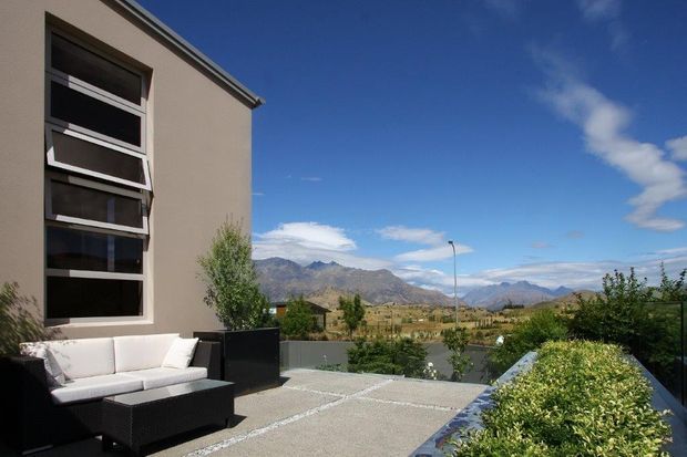 Contemporary outdoor terrace beside a modern rendered building, featuring a large dark-framed vertical window, outdoor wicker seating, paved flooring, raised planted beds, and expansive mountain views under a clear blue sky.