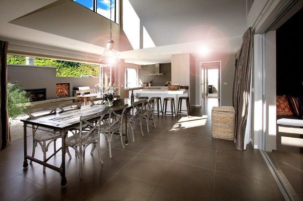 Bright open-plan interior combining a dining area, kitchen and living space. A long timber dining table with cross-back chairs sits in the foreground, while a kitchen island with bar stools is positioned behind it. The room has dark large-format tiled flooring, light walls and cabinetry, expansive glazing, a high angled ceiling, pendant lights, and a fireplace in the living area. Sliding glass doors open toward an outdoor area, creating a spacious indoor-outdoor connection.