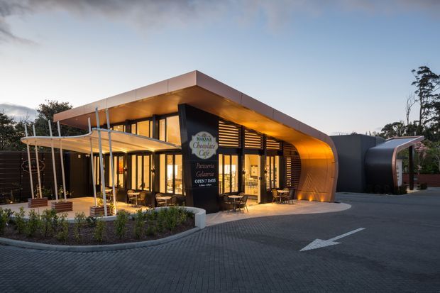 Exterior architectural photograph of a contemporary commercial chocolate and dessert venue at dusk. The building features a broad curved canopy and roofline, warm illuminated timber-toned soffits, extensive floor-to-ceiling glazing, dark vertical facade elements, a covered outdoor seating terrace, and a paved driveway with directional markings. A rounded enclosed volume extends from the main building on the right.