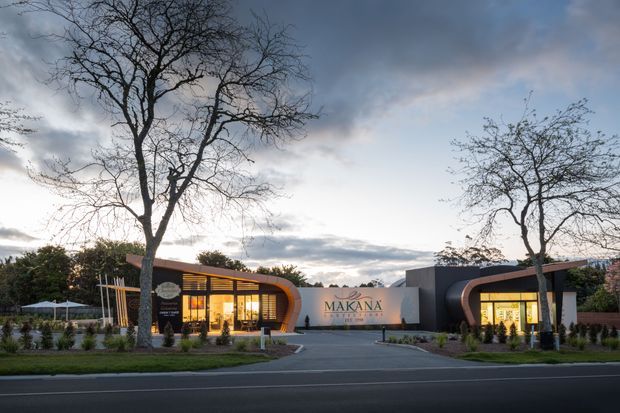 Twilight exterior view of a contemporary low-rise commercial building with two dark-clad wings, curved timber-toned architectural portals, extensive illuminated glazing, landscaped planting, and a central entrance wall marked “Makana.” The property fronts a paved roadway and is framed by mature deciduous trees.