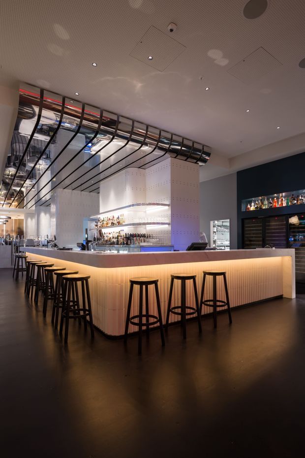 Interior photograph of a contemporary bar or hospitality venue, centered on a large illuminated bar counter with black stools. The space features dark polished flooring, white perforated bar cabinetry and walls, extensive bottle displays, concealed warm lighting, and a distinctive suspended black metal and glass ceiling installation. The atmosphere is sophisticated, dramatic, and warmly lit.